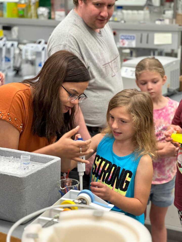 A woman shows young students a lab technique.